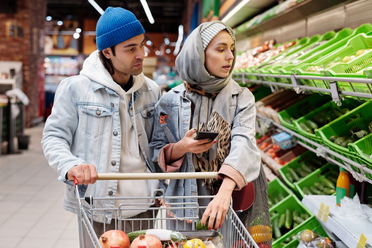 A couple pushes a cart down a supermarket aisle while one of them checks a phone.