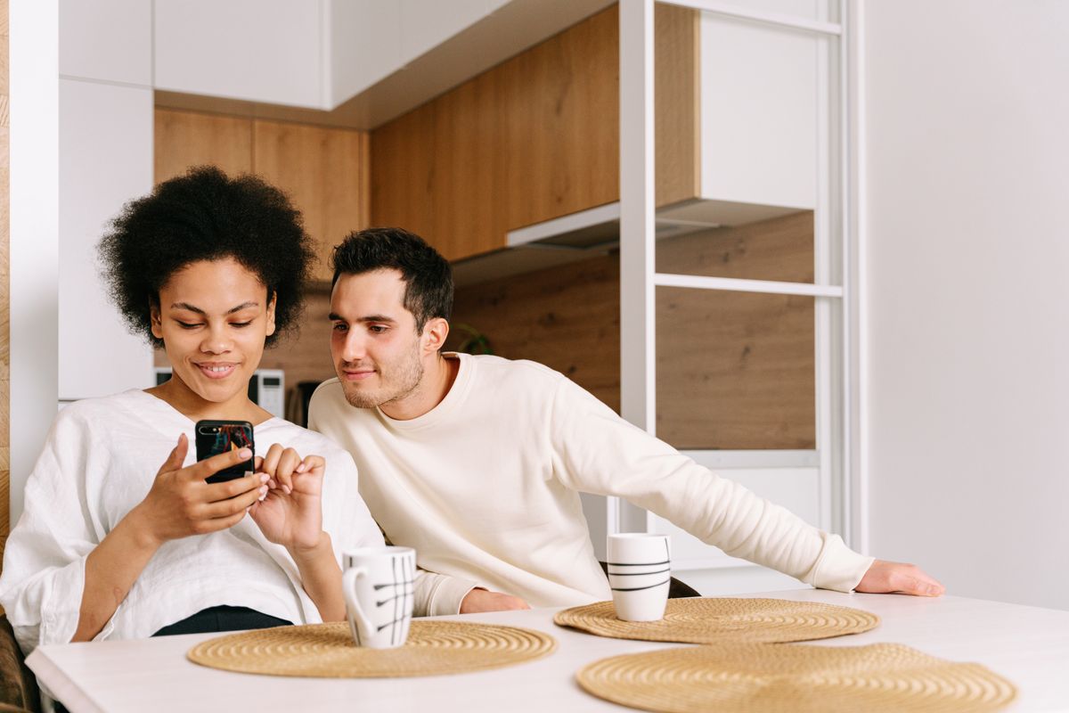 A couple sits at a kitchen table, smiling at a phone together.