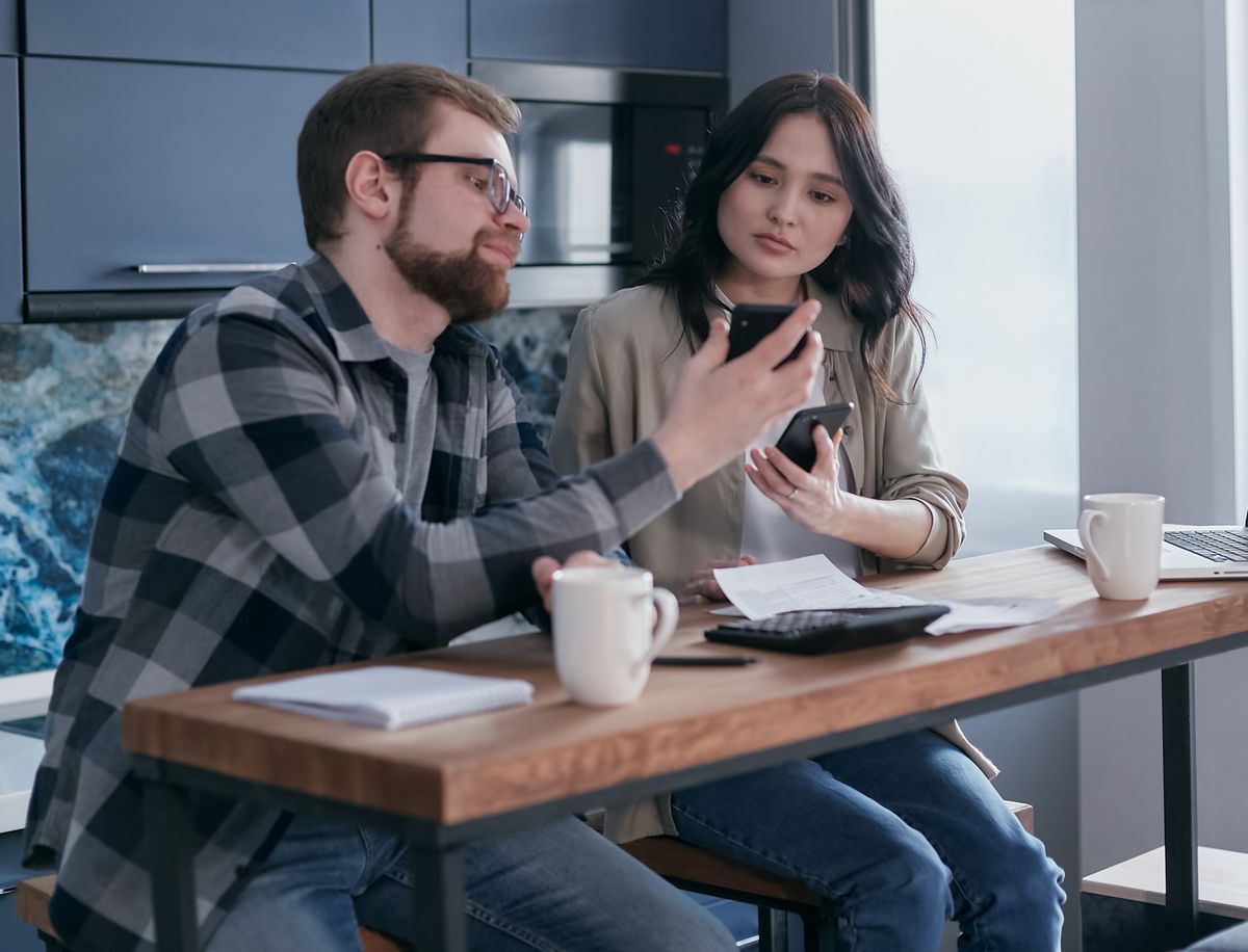 A couple sit at a table with a laptop, phones and coffee, talking through their plans.
